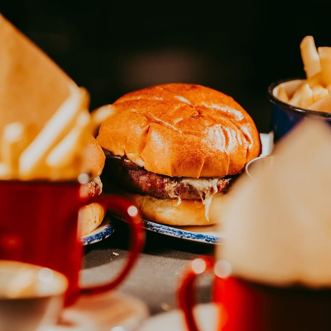 Juicy burger with melted cheese on a toasted bun, surrounded by red cups and golden fries in a cozy dining atmosphere.