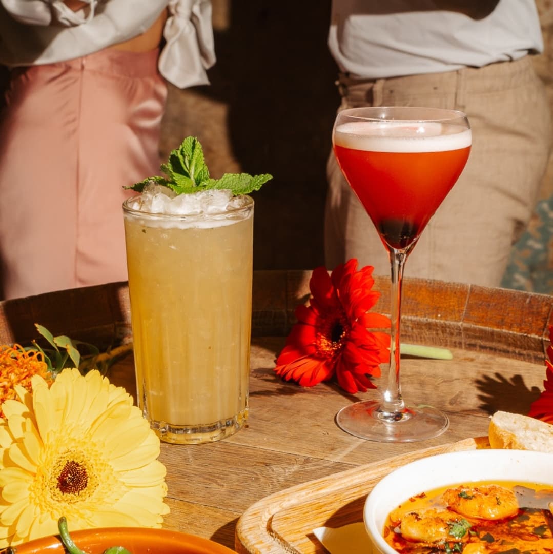 Two vibrant cocktails on a wooden tray, surrounded by colorful flowers and a dish of shrimp, creating a lively dining atmosphere.