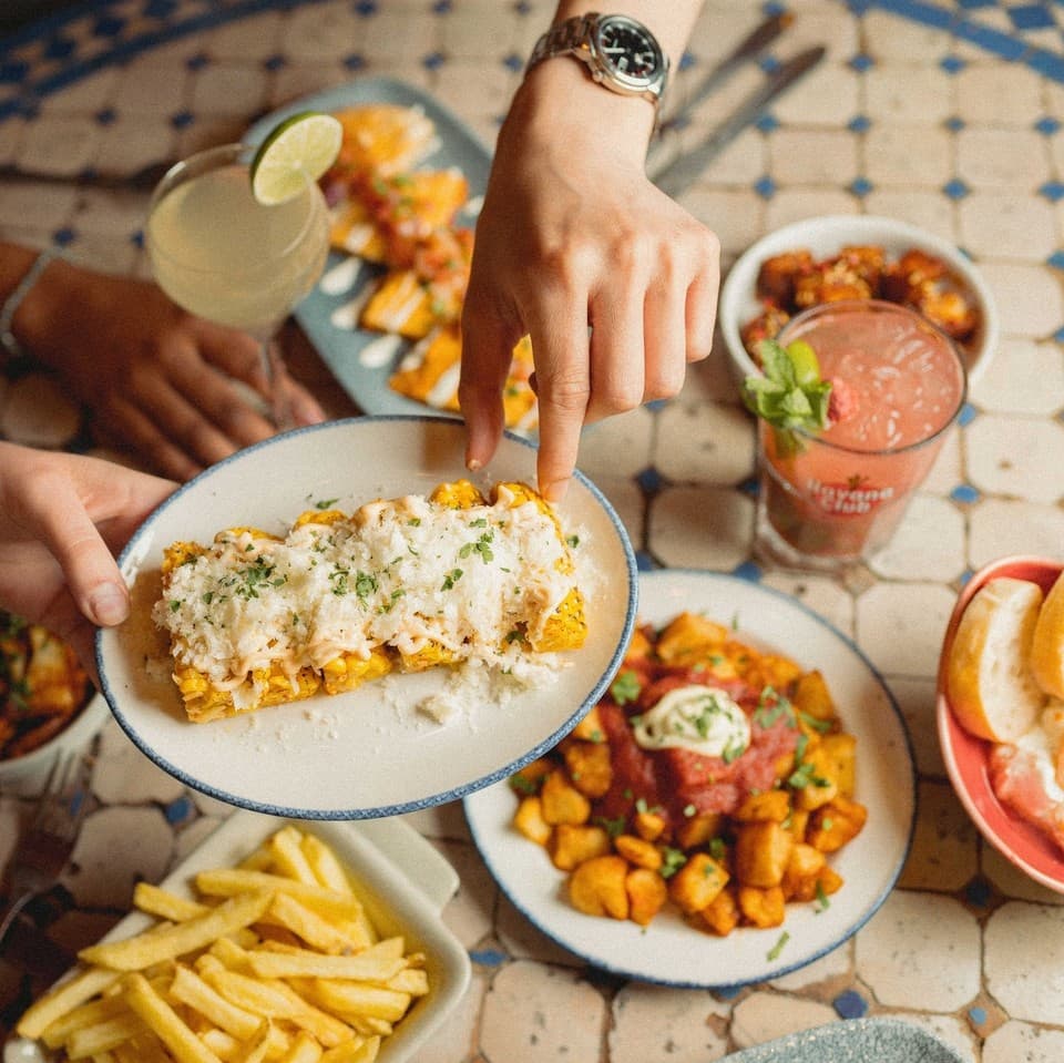 A hand reaches for a plate of vibrant tacos, surrounded by colorful cocktails and sides of fries and salad on a tiled table.