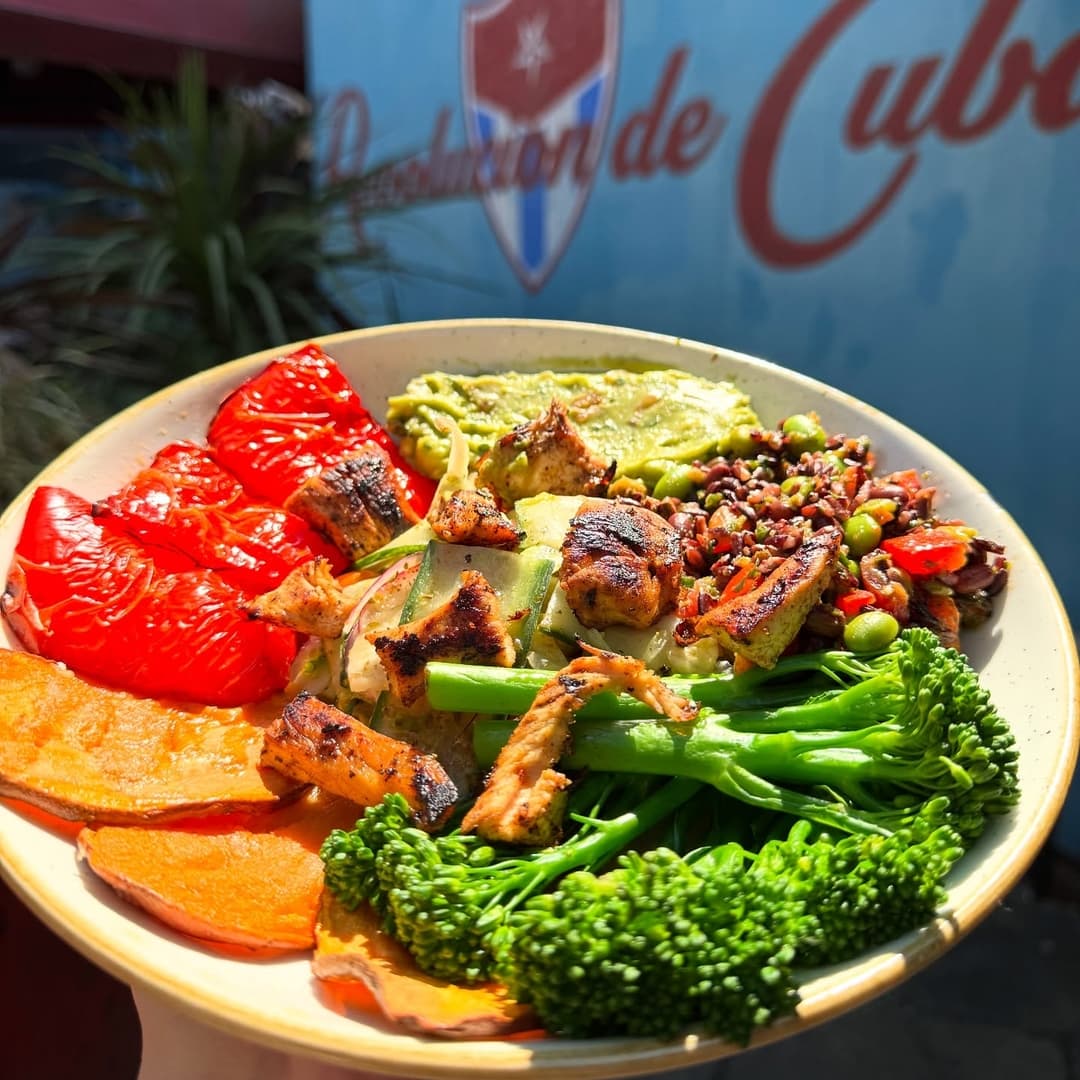 Vibrant plate of grilled chicken, colorful vegetables, quinoa, and guacamole against a backdrop of Revolución de Cuba sign.