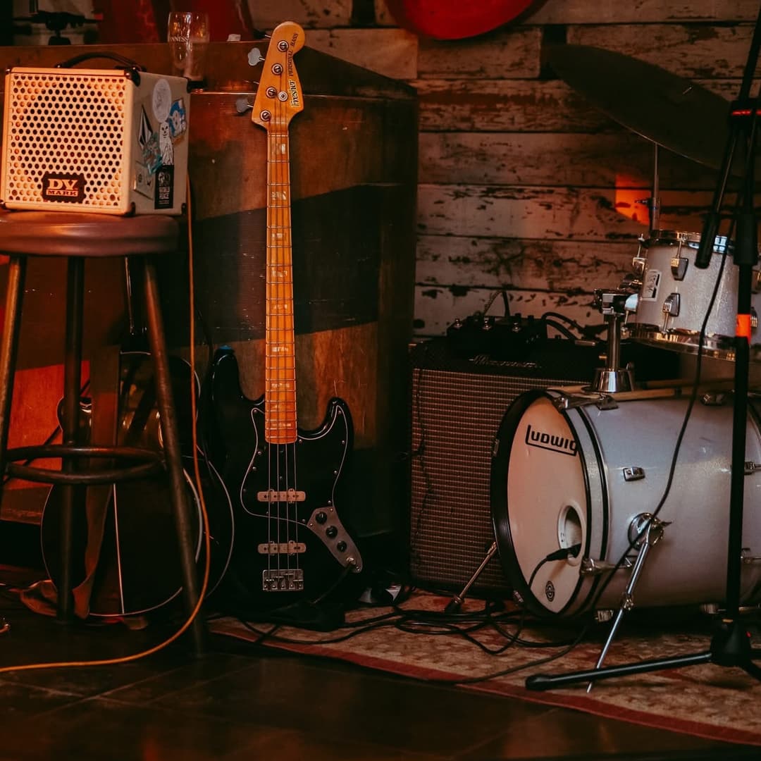 Warmly lit corner of a vibrant venue featuring a bass guitar, drum set, and a stool, creating a lively atmosphere.