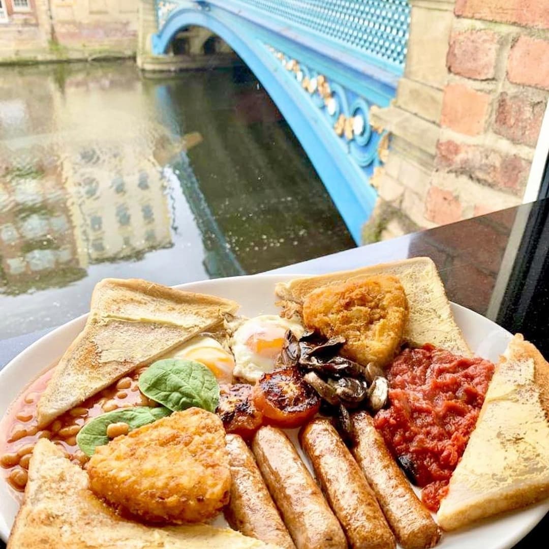 A hearty breakfast plate with toast, sausages, beans, and tomatoes, overlooking a blue bridge and calm water.