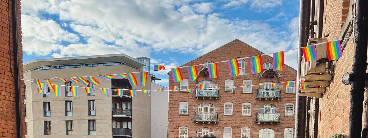 Colorful rainbow flags strung between buildings under a bright blue sky with fluffy clouds, creating a festive atmosphere.