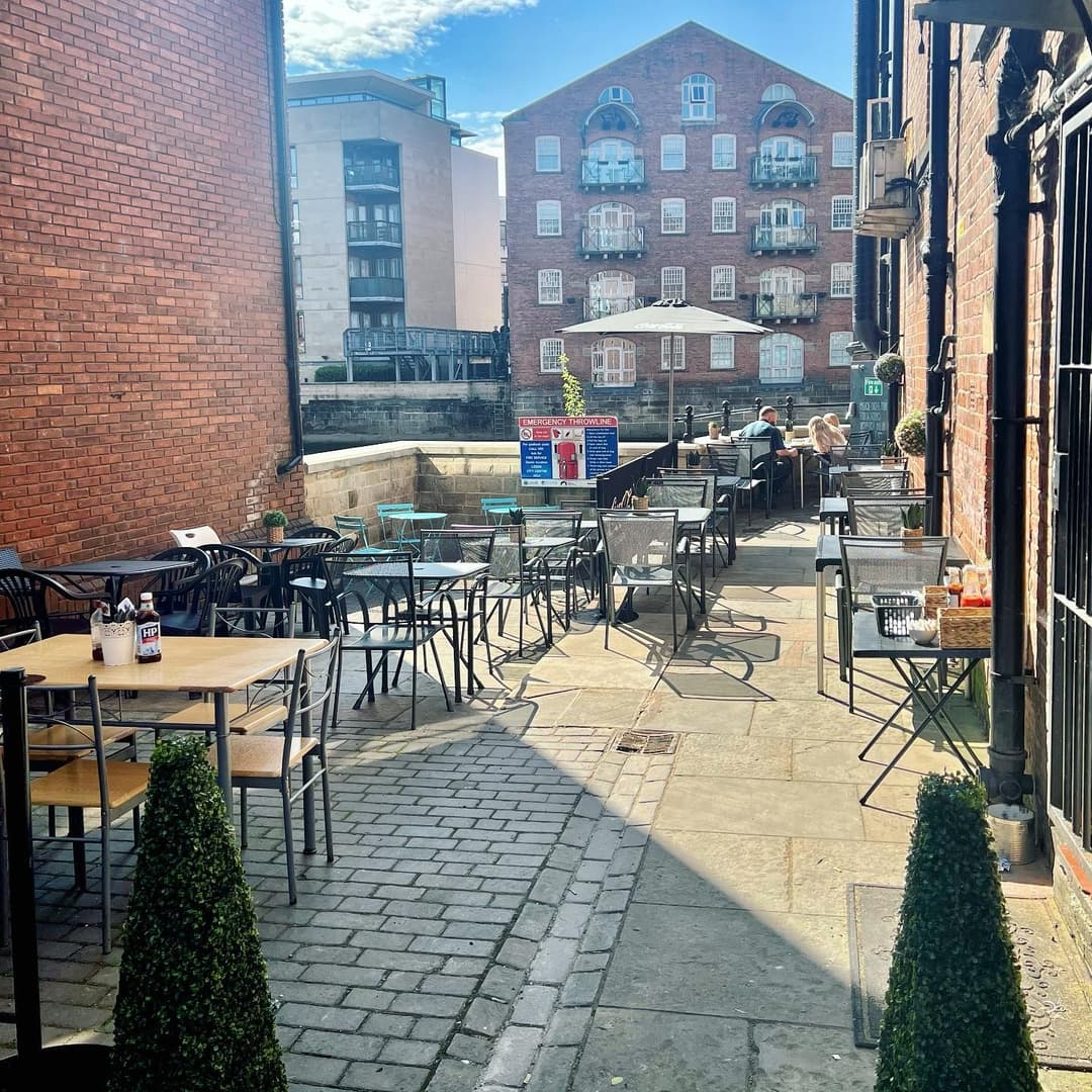 Charming outdoor dining area with tables, potted plants, and a view of the river, set against historic brick buildings.