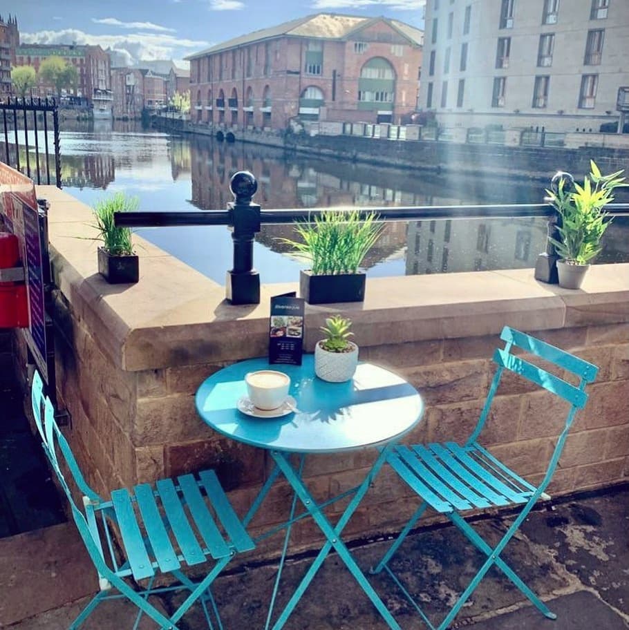 Bright blue table and chairs by a serene river, adorned with potted plants, creating a cozy outdoor dining atmosphere.
