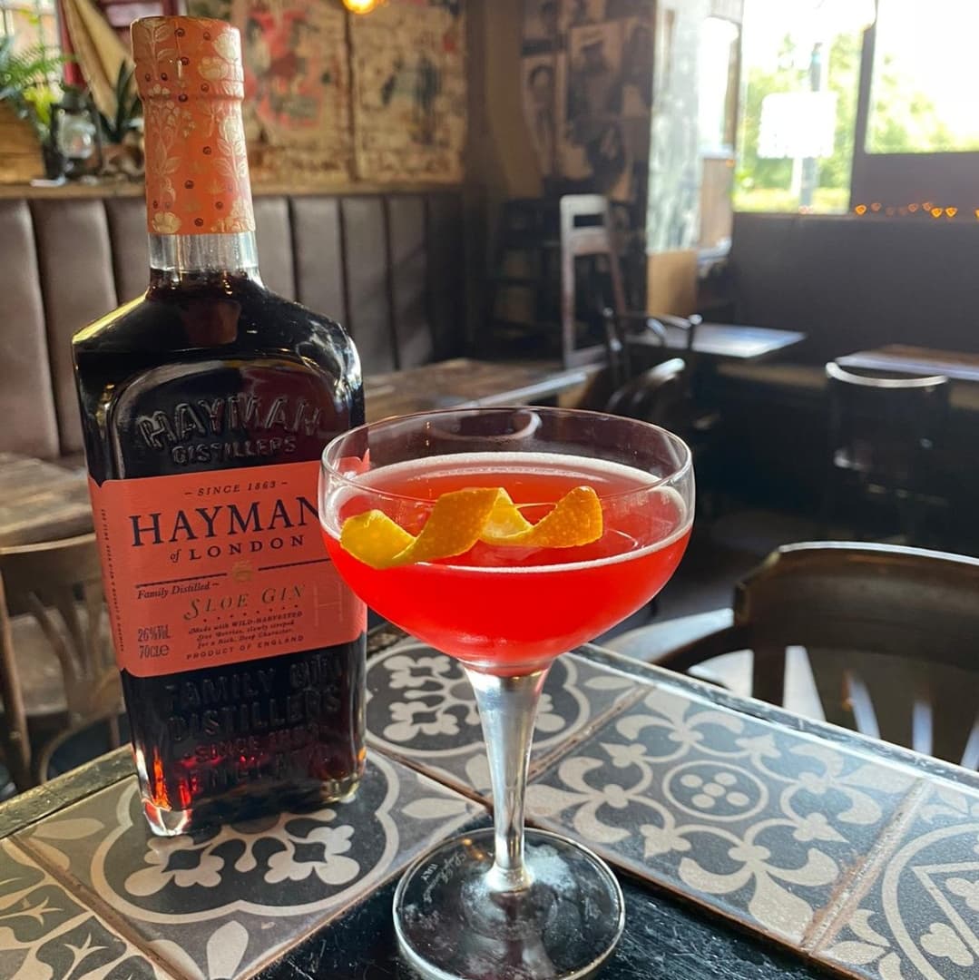 A vibrant cocktail garnished with citrus peels beside a bottle of Hayman’s gin, set in a cozy, rustic bar.