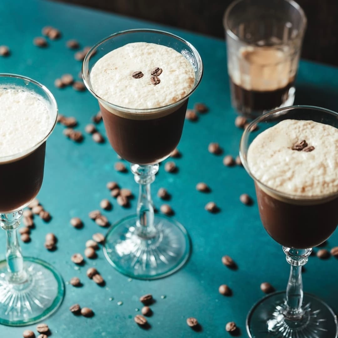 Three elegant glasses of coffee cocktails topped with frothy cream and coffee beans, surrounded by scattered coffee beans on a teal surface.