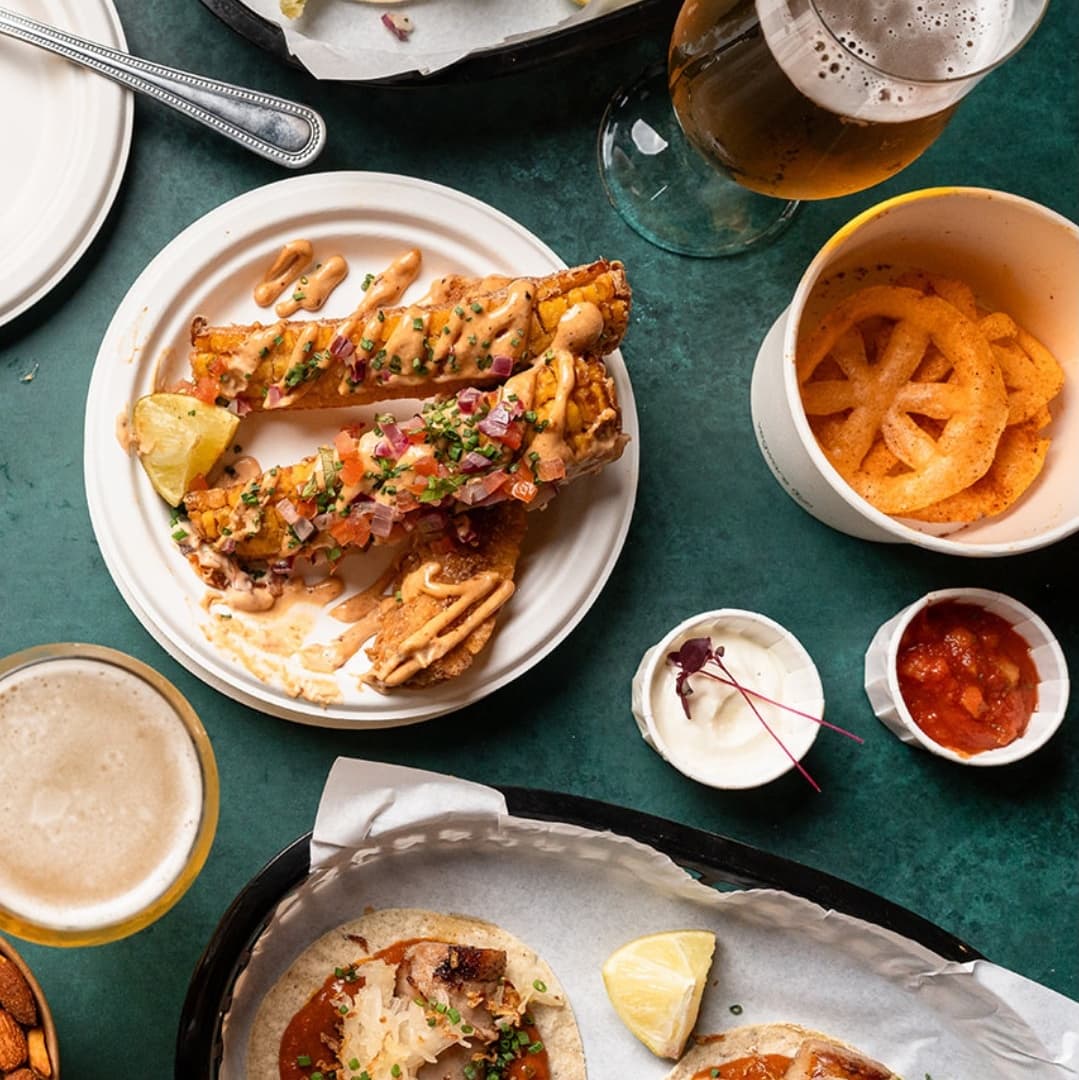 Colorful plate of tacos with toppings, crispy snacks, and drinks on a vibrant green table, creating a lively dining atmosphere.