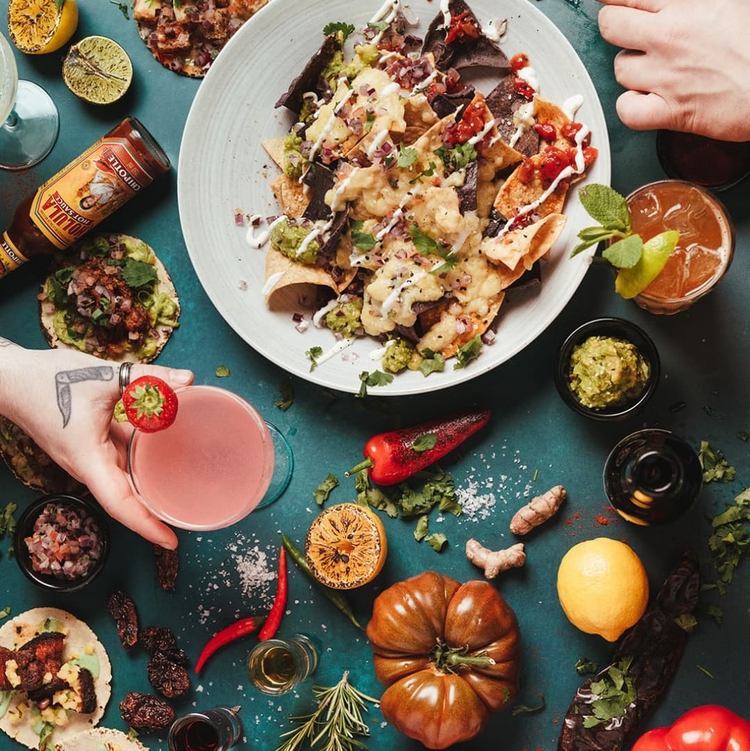 Colorful spread of nachos topped with guacamole, salsa, and cheese, surrounded by tacos, fresh ingredients, and cocktails.
