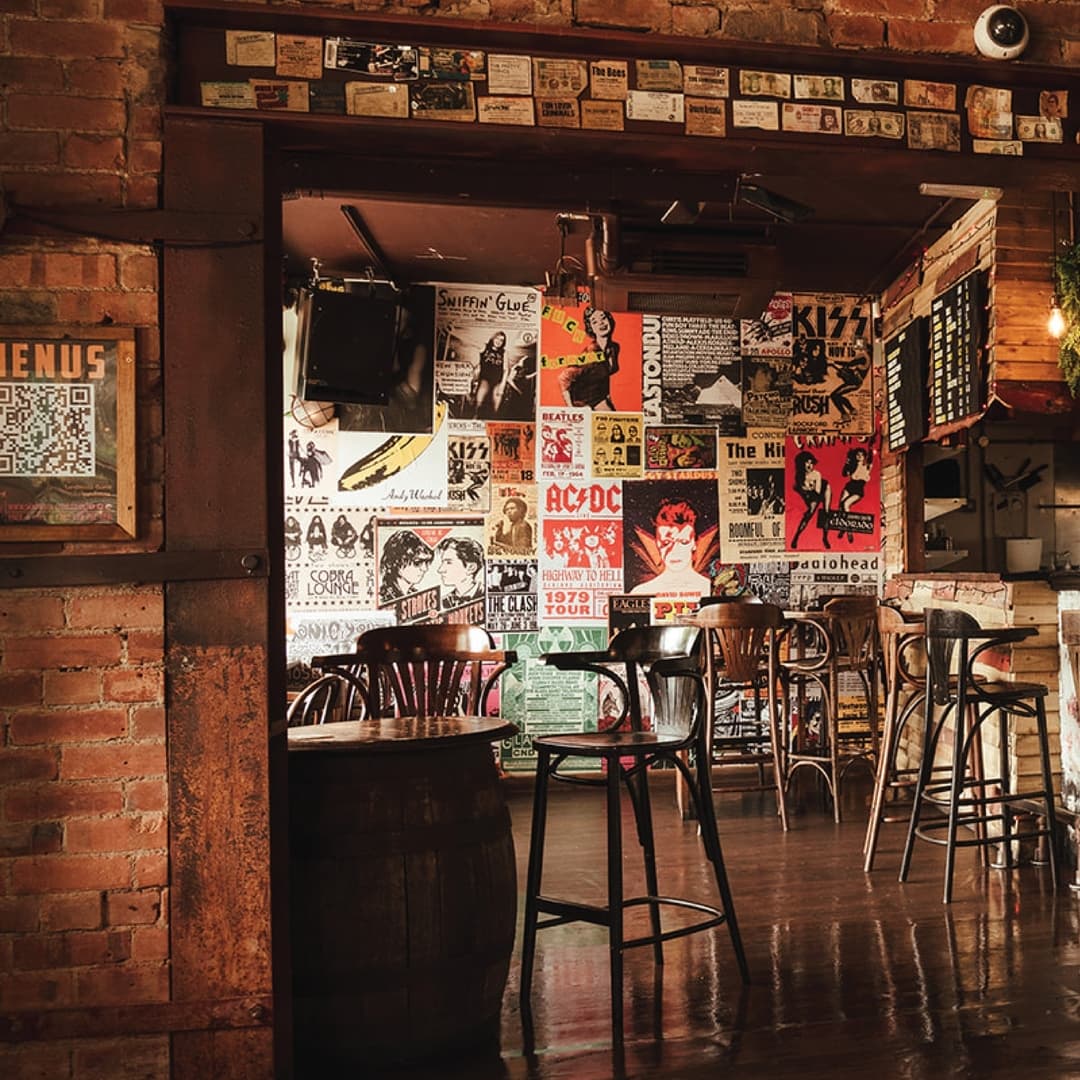 Cozy bar interior with wooden chairs, exposed brick walls adorned with vibrant music posters, and warm lighting.