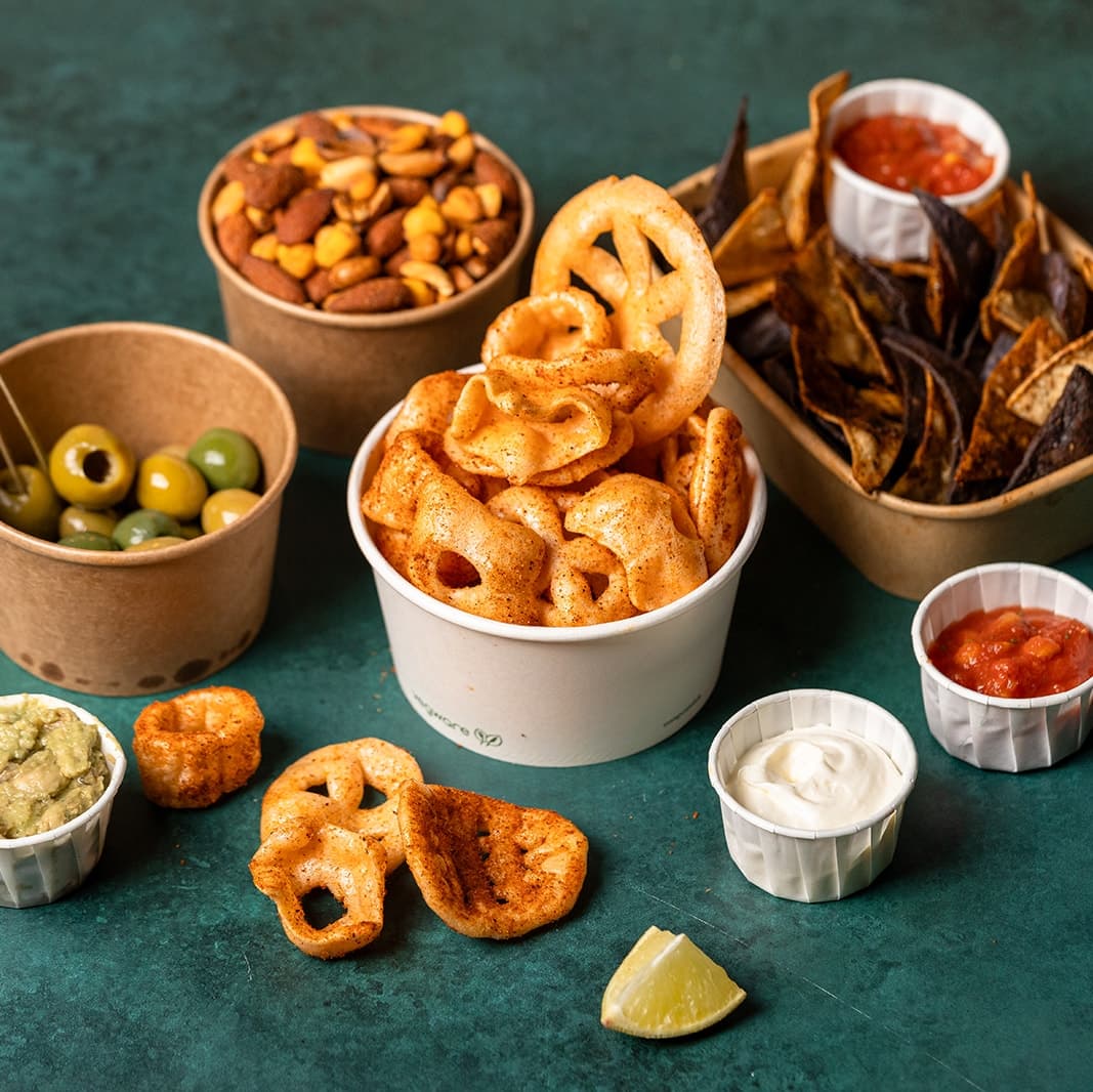 Colorful bowls of snacks including crispy chips, nuts, olives, and dips, arranged on a vibrant green table.