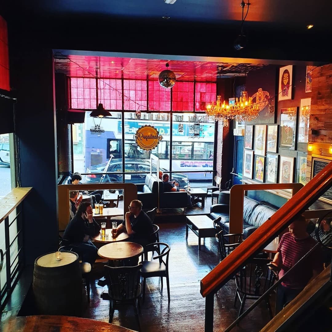 Cozy bar scene with colorful decor, patrons enjoying drinks, and a warm atmosphere enhanced by a chandelier and art on the walls.