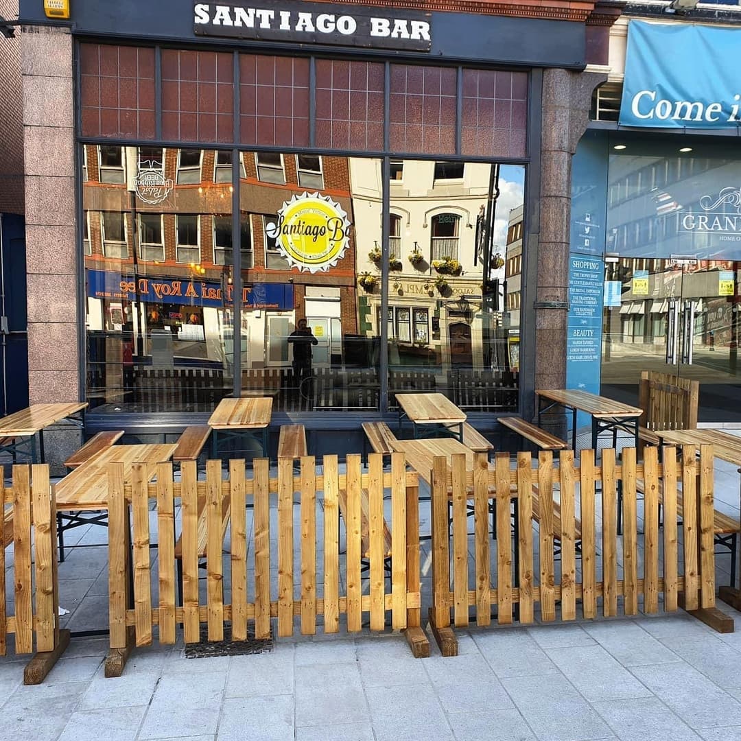 Outdoor seating area at Santiago Bar with wooden tables and a rustic fence, inviting atmosphere with a city backdrop.