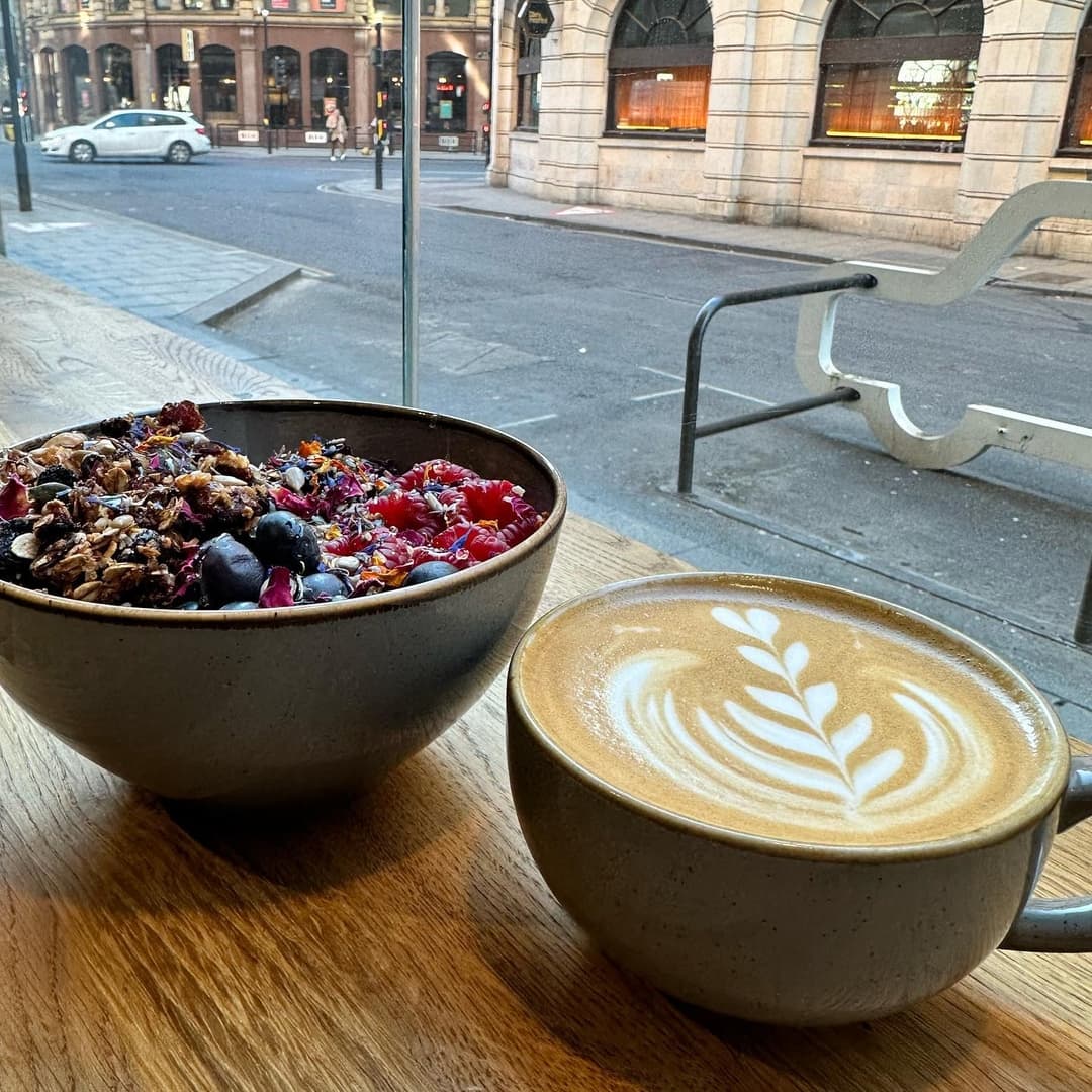 A cozy café scene featuring a bowl of colorful granola topped with berries and a latte with a leaf design.
