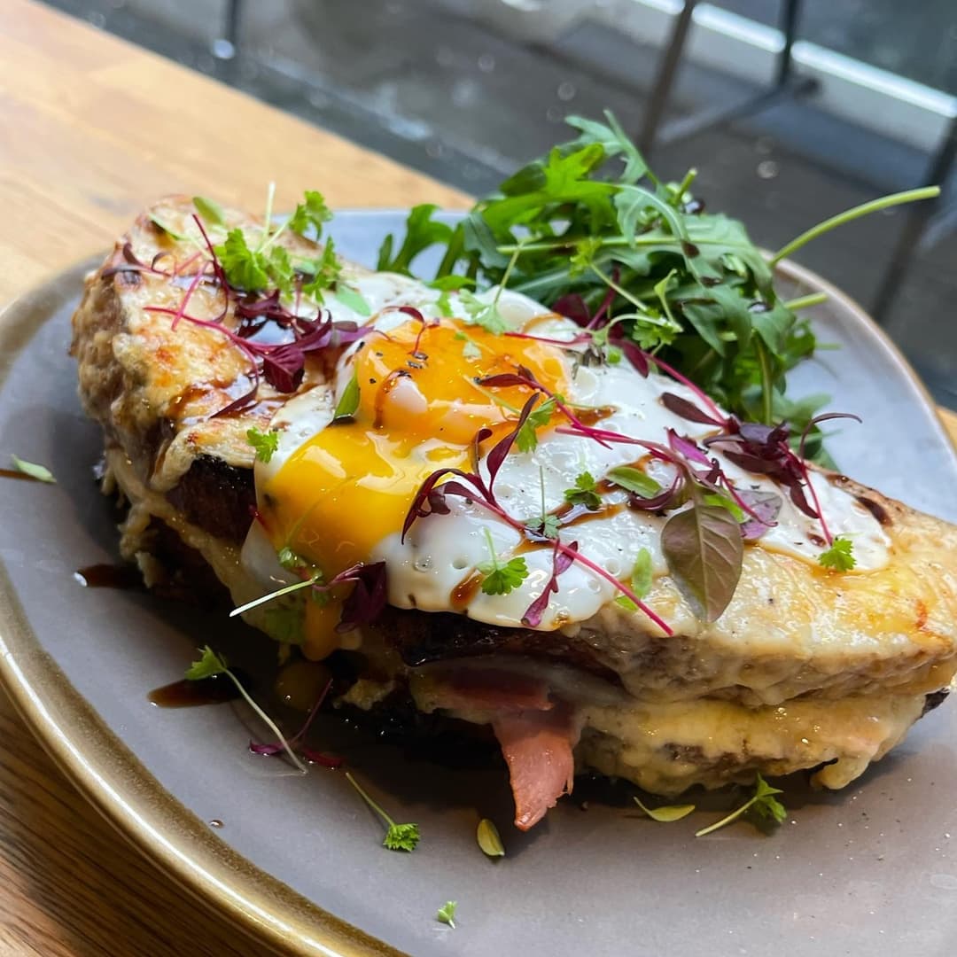 A gourmet dish featuring a toasted sandwich topped with melted cheese, a runny egg, and fresh herbs, served with greens.