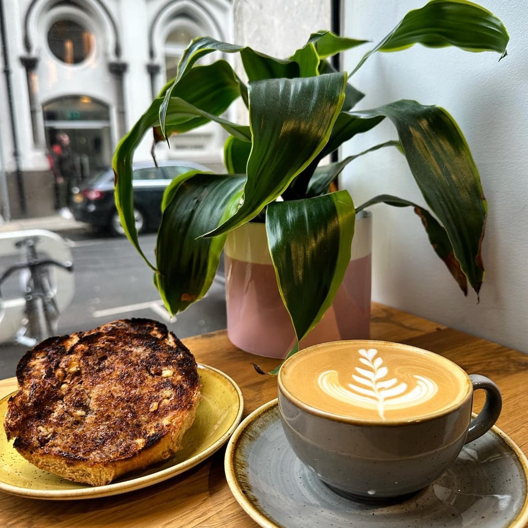 Warm latte with leaf design beside toasted bread on a yellow plate, with a green plant and city view in the background.
