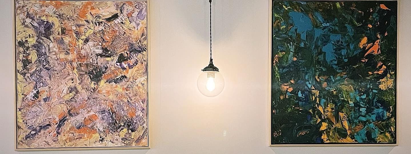 Two vibrant abstract paintings hang on a wall, illuminated by a warm pendant light, creating a cozy atmosphere.