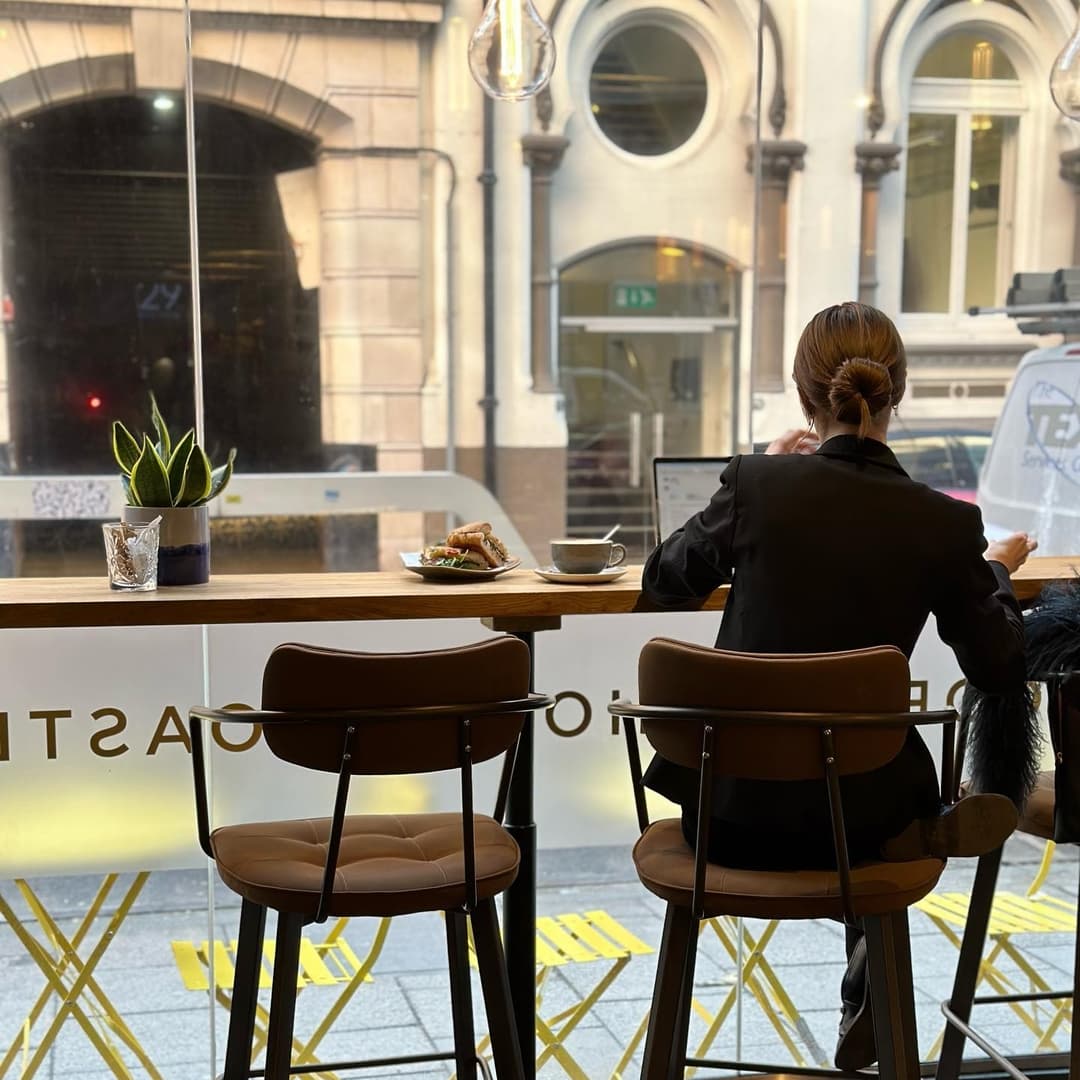 A woman in a black suit sits at a wooden counter, enjoying coffee and a sandwich, with a view of the street outside.
