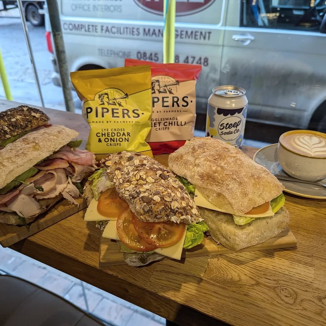 Freshly made sandwiches with assorted fillings, chips, and a latte on a wooden table, with a cozy café atmosphere.