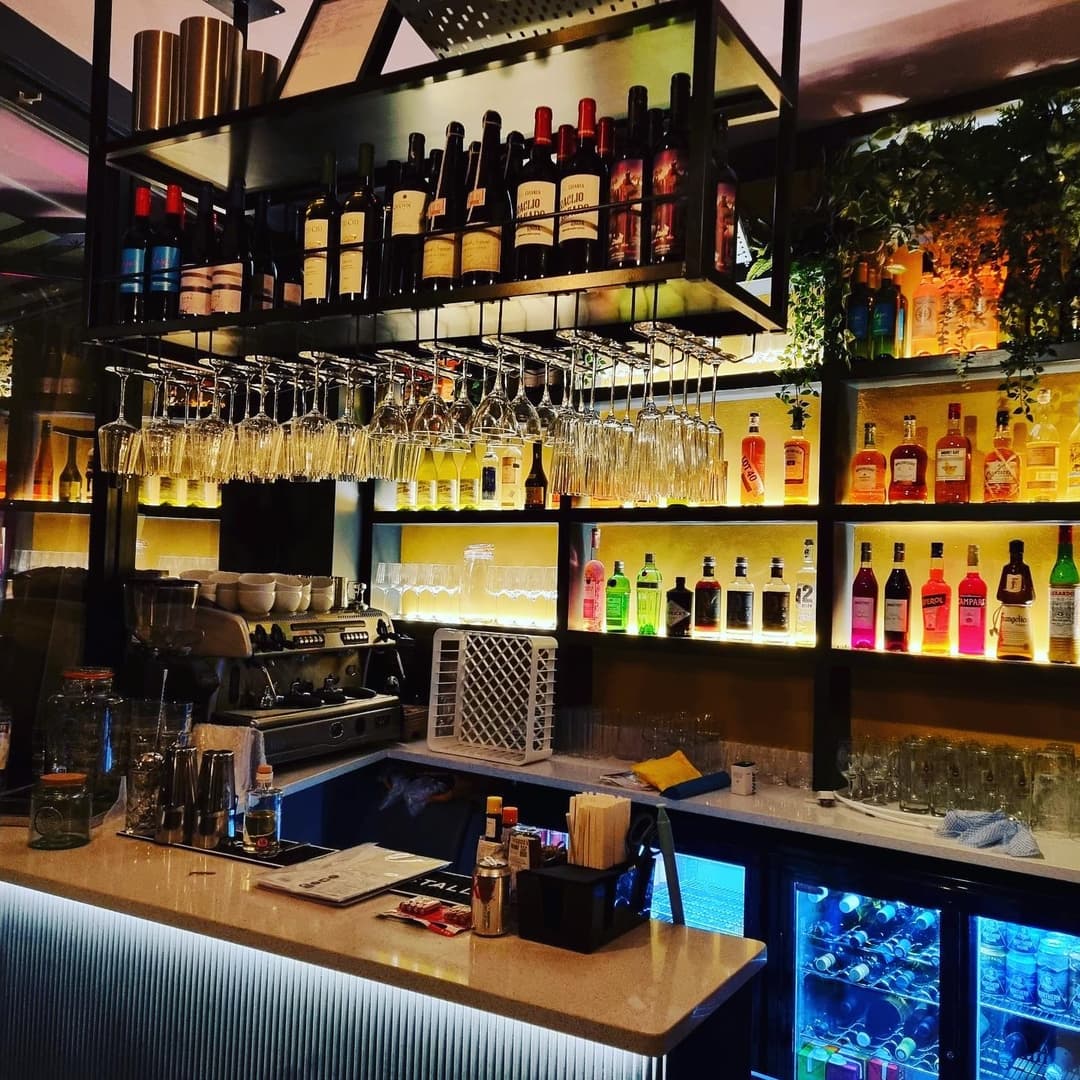 Stylish bar with a sleek countertop, illuminated shelves of colorful bottles, and hanging wine glasses, creating a vibrant atmosphere.
