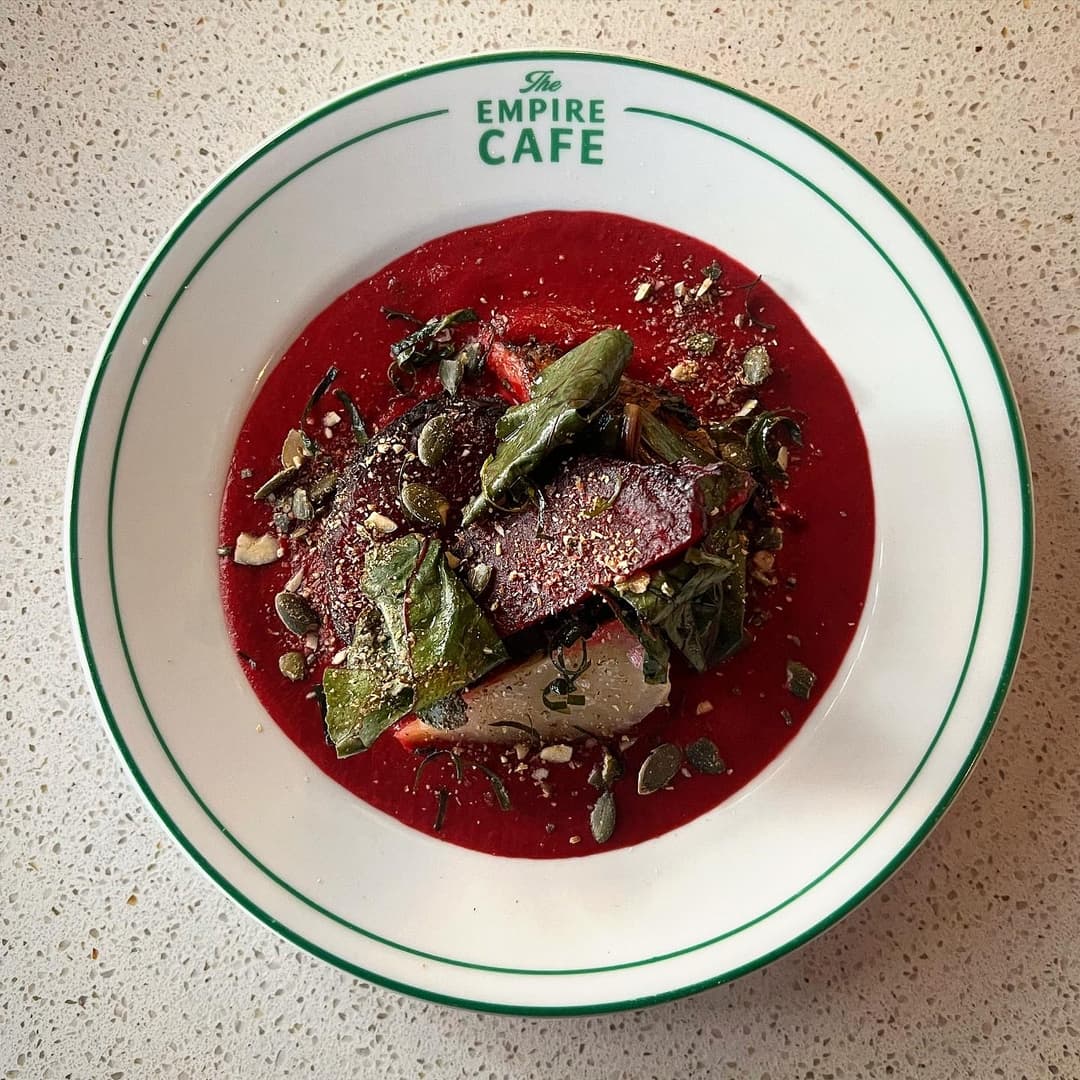 Vibrant beet soup topped with greens, seeds, and herbs, served in a classic white bowl with green rim at The Empire Cafe.