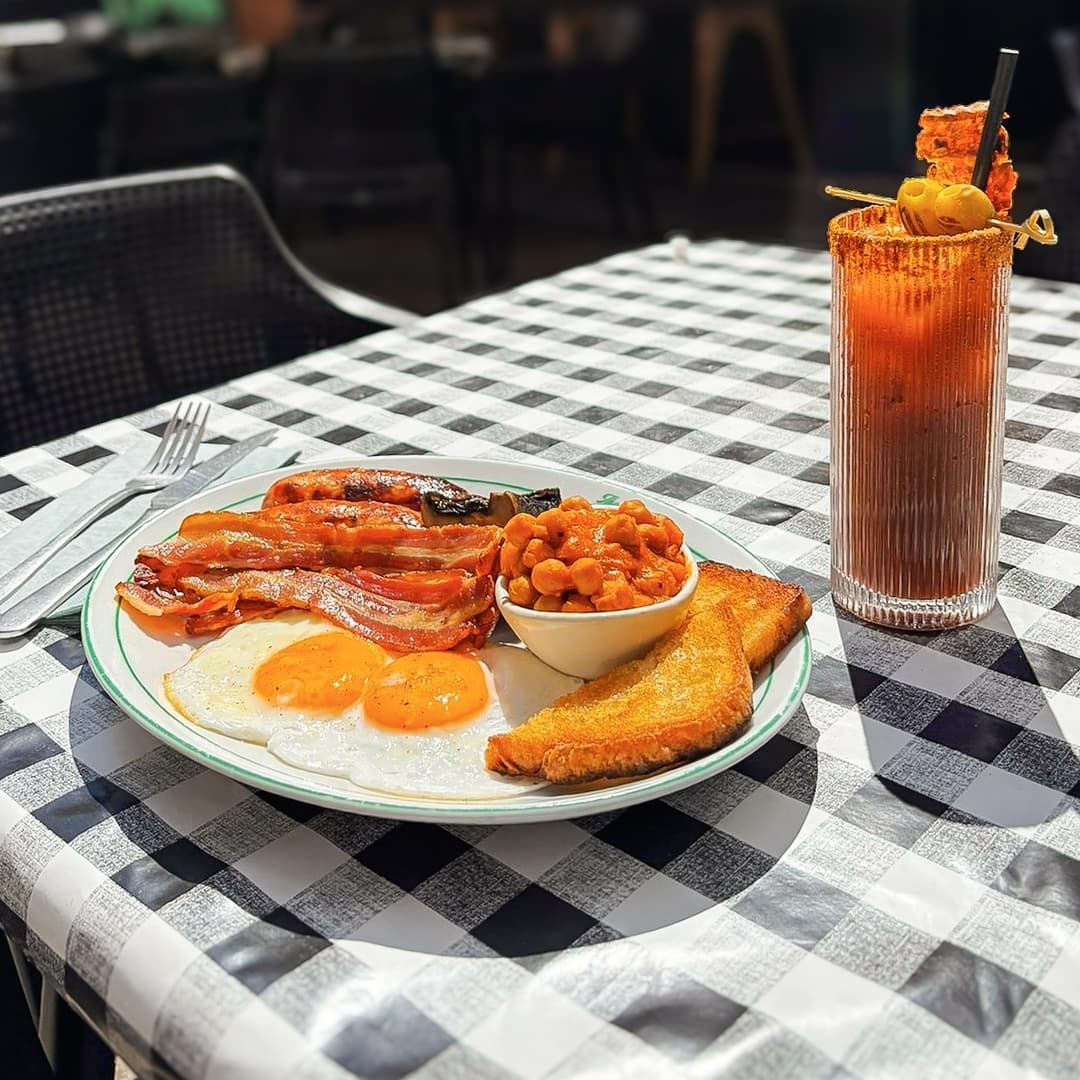 A hearty breakfast plate with eggs, bacon, baked beans, and toast beside a tall, garnished Bloody Mary on a checkered tablecloth.