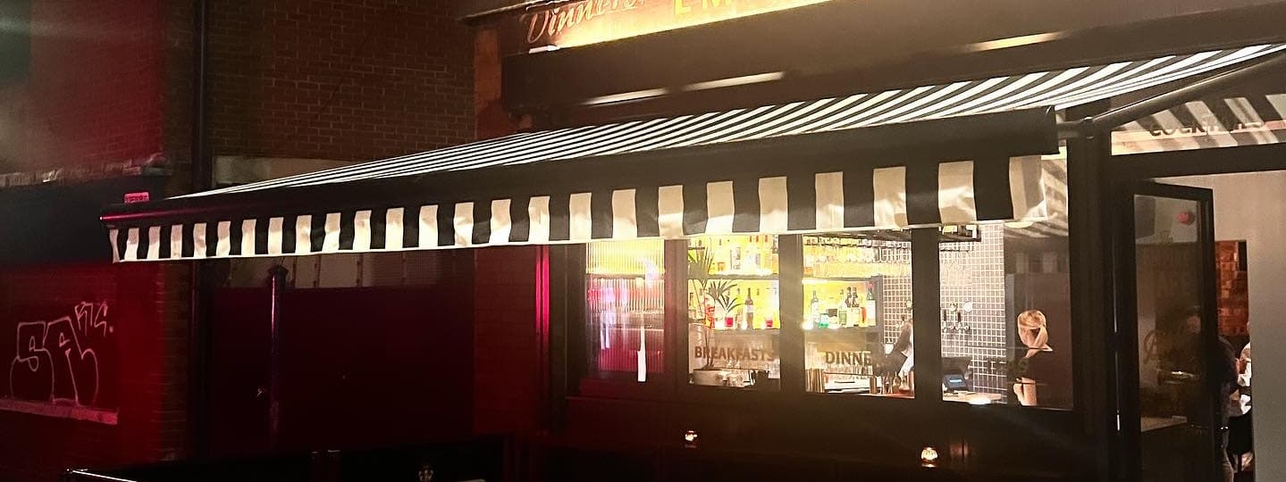Charming café exterior with a striped awning, warm lighting, and a glimpse of a vibrant bar scene inside.