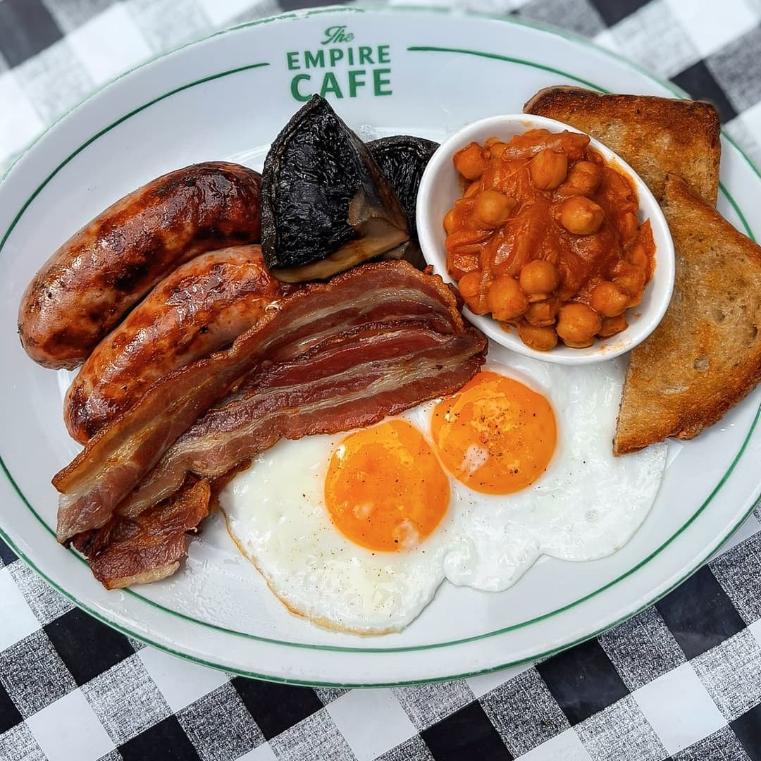 A hearty breakfast plate featuring sausages, bacon, eggs, baked beans, black pudding, and toast on a checkered tablecloth.