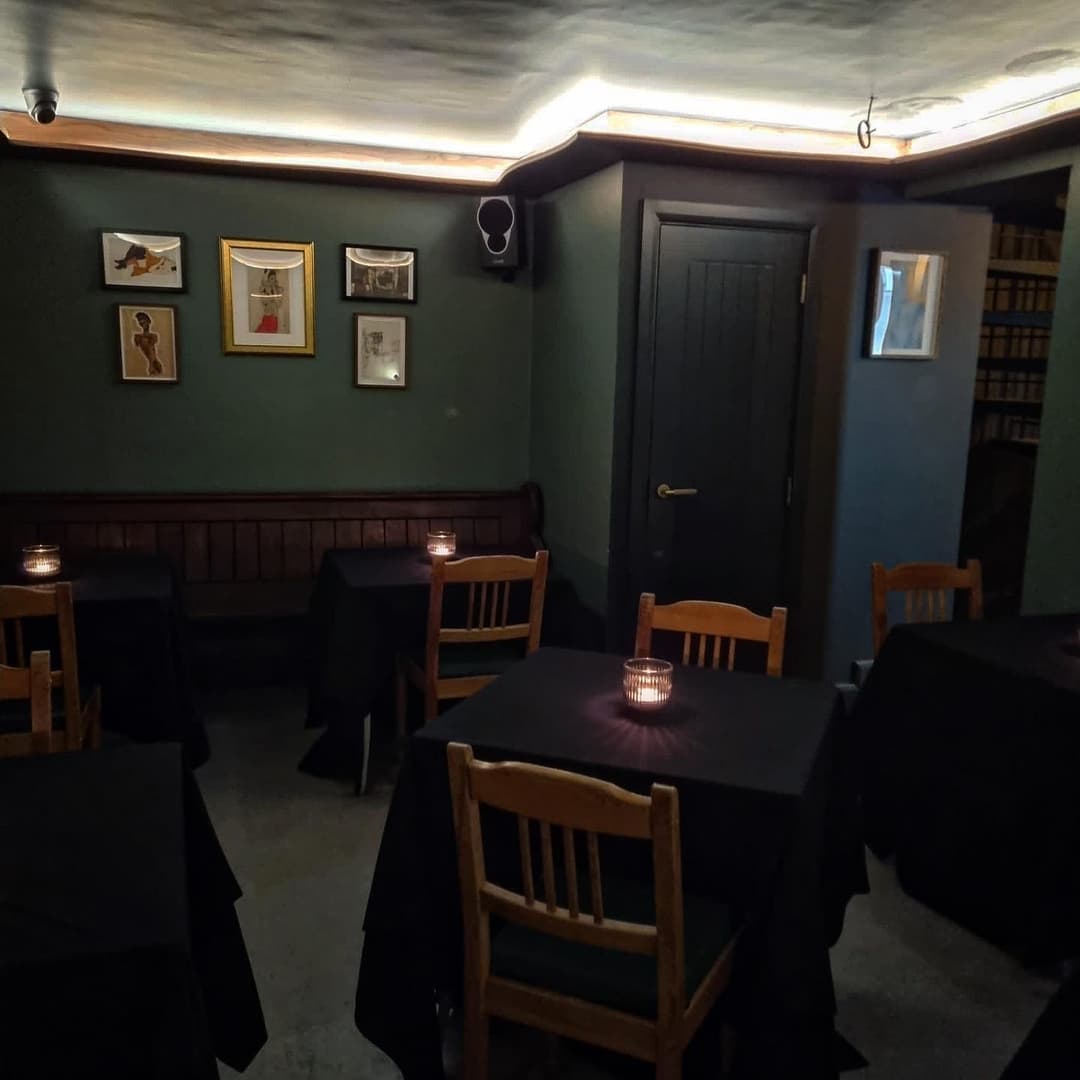 Cozy dining area with dark tables, flickering candles, and framed artwork on green walls, creating an intimate atmosphere.