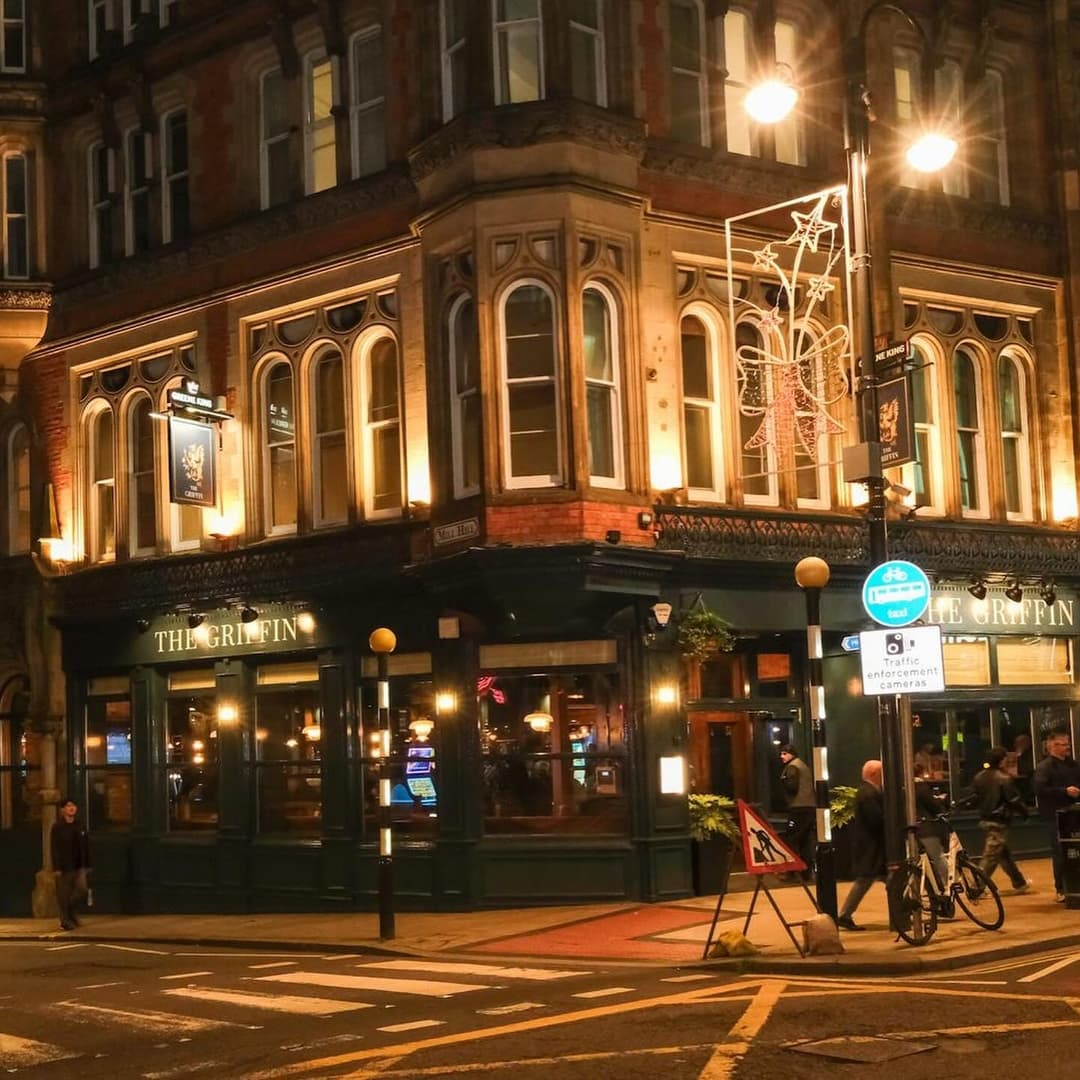 The Griffin, a warmly lit corner pub, features large windows and festive decorations, with people gathered outside.