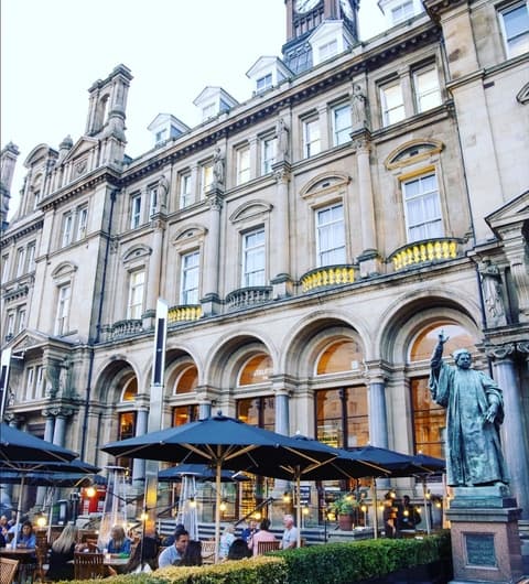 Elegant outdoor dining area with umbrellas, historic building facade, and a statue, creating a vibrant atmosphere.
