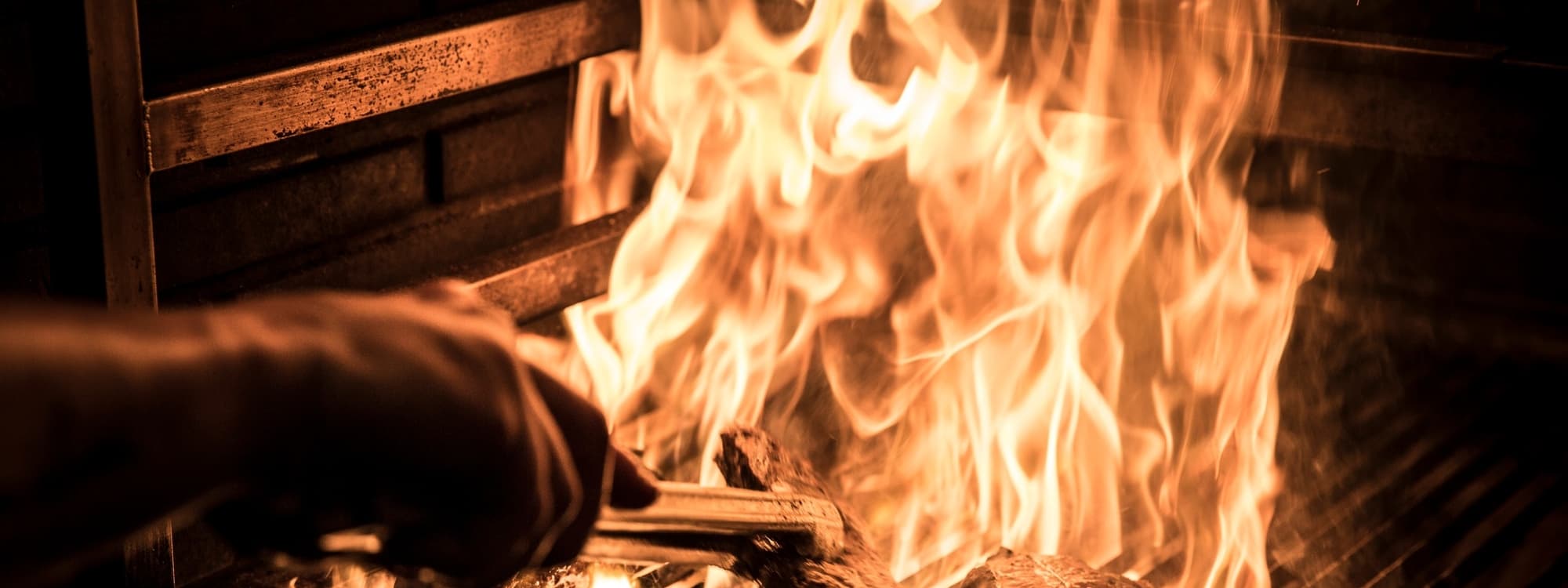 Flames dance in a grill, with a hand using a spatula to manage the sizzling fire, creating a warm, inviting atmosphere.