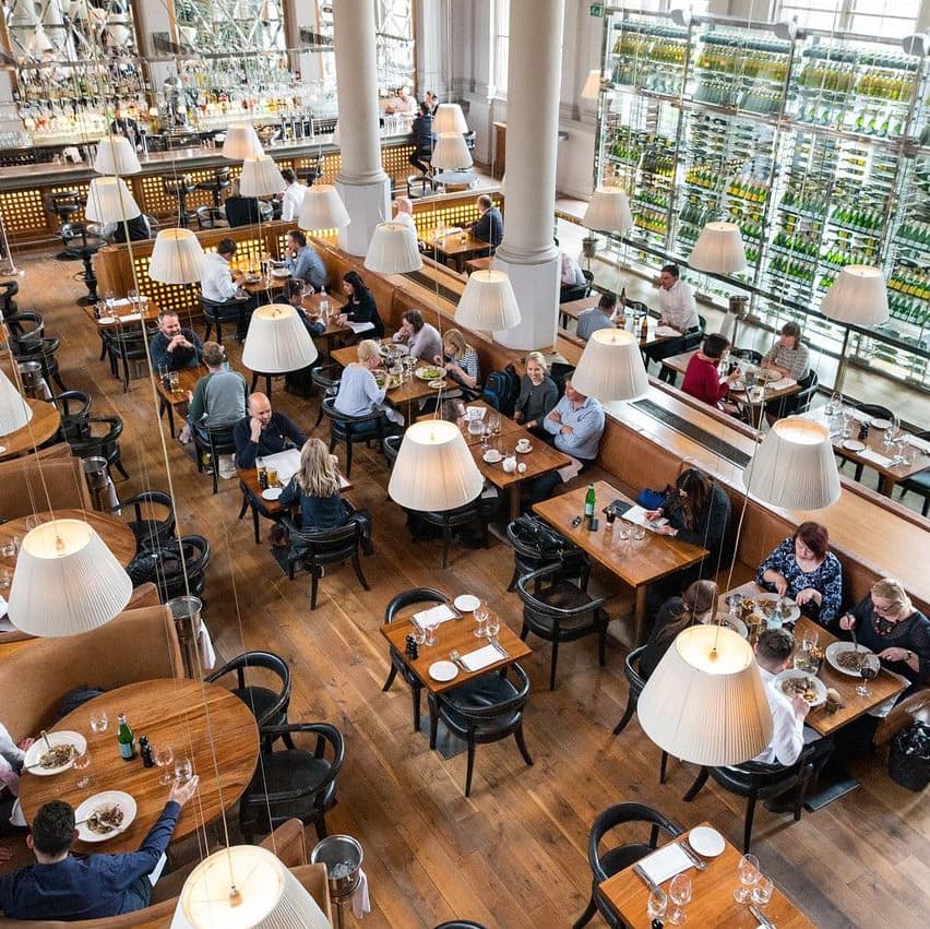 Spacious restaurant with wooden floors, large windows, and elegant lighting; diners enjoy meals at stylish tables.