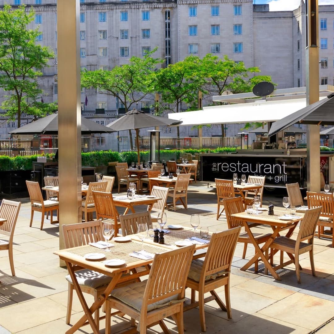 Sunny outdoor dining area with wooden tables and chairs, surrounded by greenery and modern architecture.