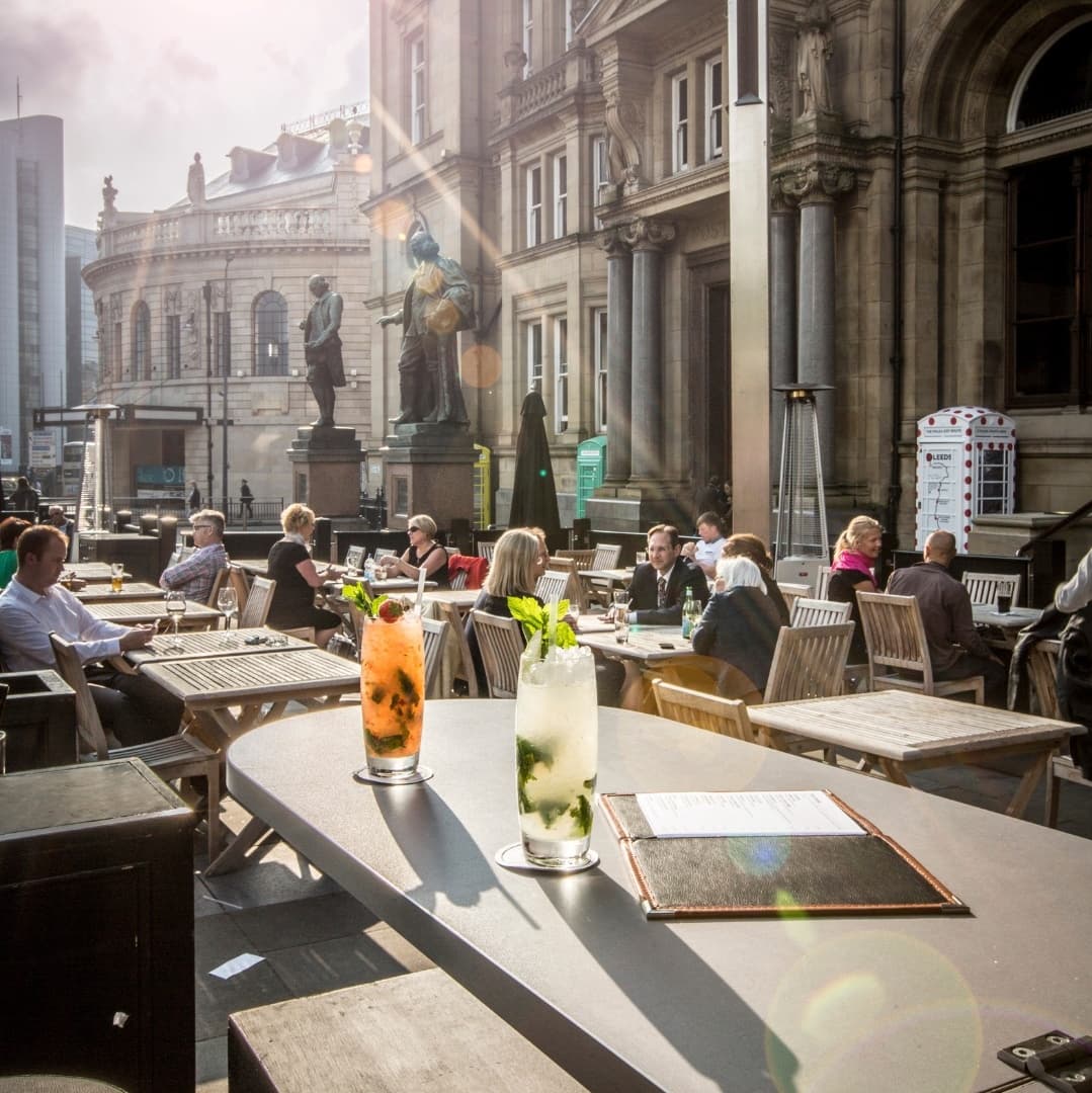 Two colorful cocktails on a table, with a bustling outdoor terrace and historic architecture in the background.