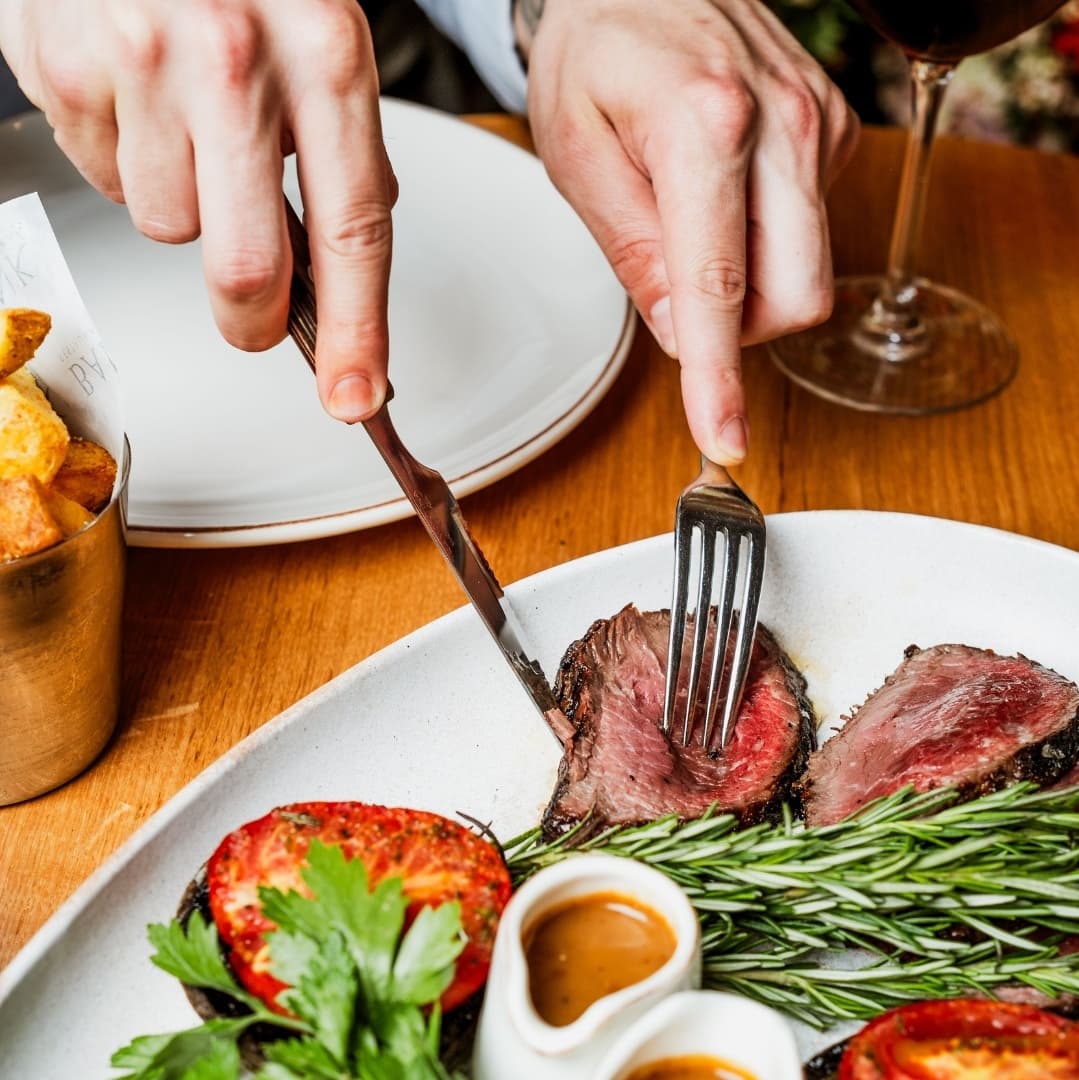 A close-up of hands cutting into a juicy steak, garnished with rosemary and tomatoes, with a glass of red wine nearby.