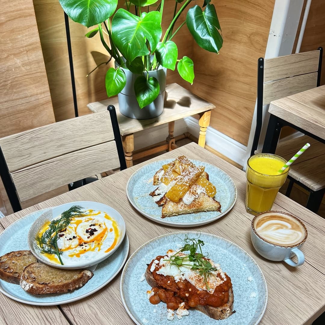 Brightly lit table with three plates of colorful dishes, a glass of orange juice, and a latte, surrounded by greenery.