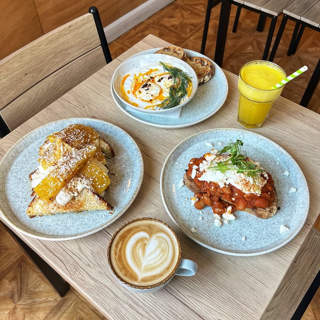 A cozy table with a latte, vibrant orange juice, and three colorful dishes: toast with pineapple, yogurt, and a savory bean dish.