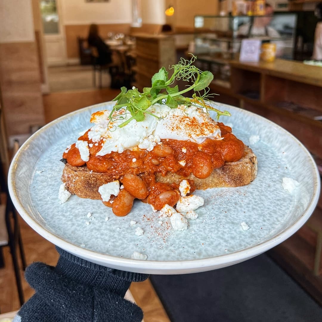 A plate of toast topped with spicy beans, poached egg, crumbled feta, and fresh greens, set in a cozy café atmosphere.