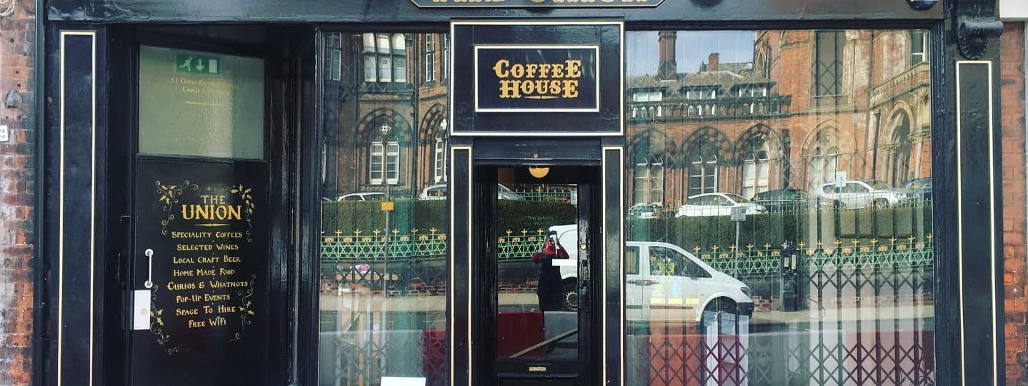 Charming café entrance with large glass windows, inviting signage, and a view of greenery and historic buildings outside.
