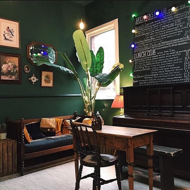 Cozy coffee house with green walls, a large plant, vintage decor, and a wooden table beside a piano.