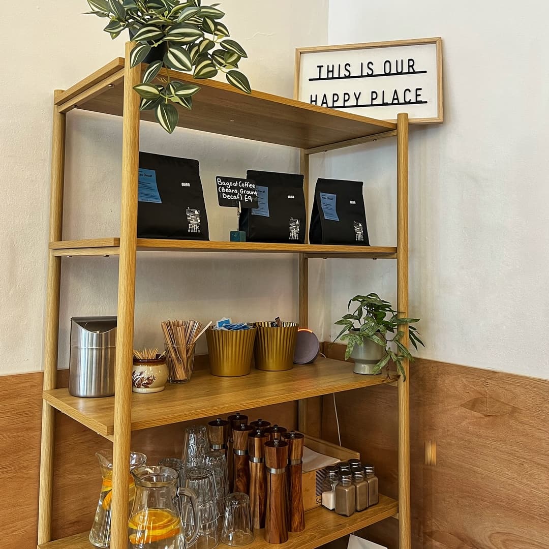 Wooden shelf with coffee bags, glassware, and plants, under a sign reading "THIS IS OUR HAPPY PLACE." Cozy atmosphere.