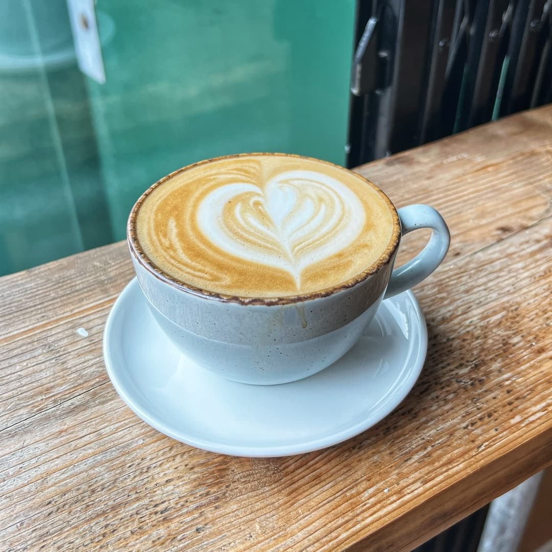 A latte with a heart-shaped foam design sits on a white saucer atop a rustic wooden table.
