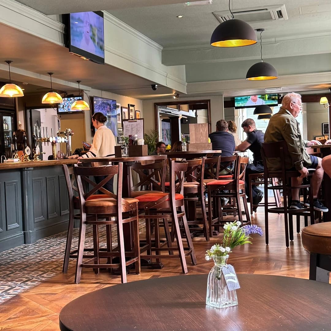 Cozy pub interior with wooden bar, patrons seated at tables, and warm lighting creating a welcoming atmosphere.