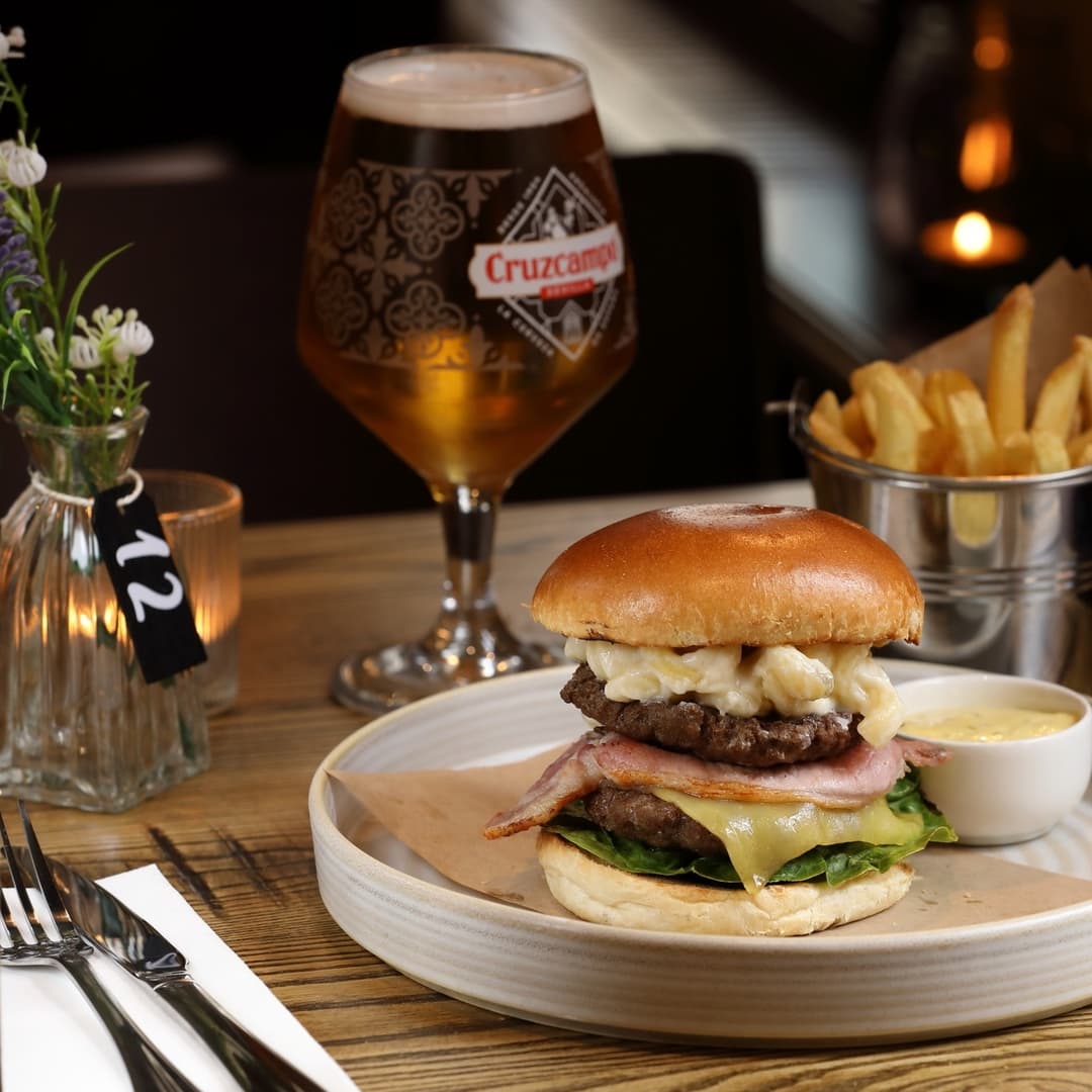 Juicy burger stacked with cheese, ham, and lettuce, served with fries and a pint of beer on a rustic wooden table.
