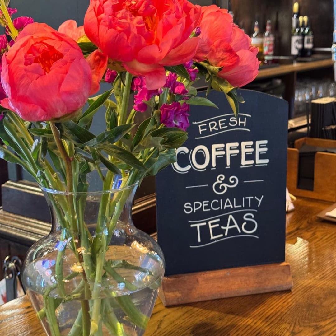Vibrant pink peonies in a vase beside a chalkboard sign reading "Fresh Coffee & Speciality Teas" in a cozy café setting.