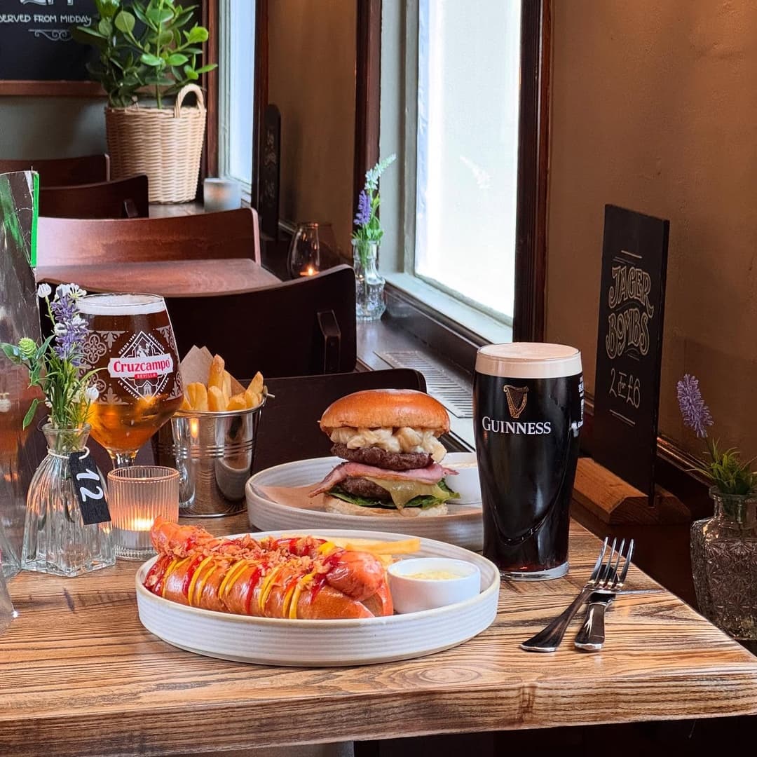 A wooden table displays a gourmet burger, hot dog with toppings, fries, and drinks, creating a cozy dining atmosphere.