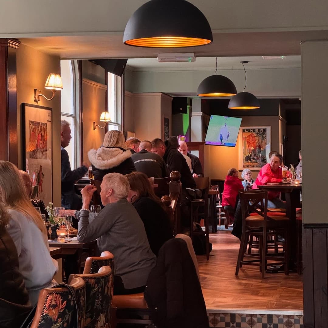 Cozy pub interior with patrons enjoying drinks, warm lighting, and a lively atmosphere, featuring wooden furnishings and artwork.