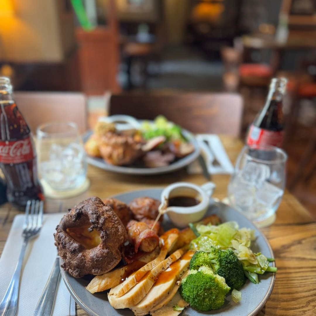 Plates of roast chicken, Yorkshire pudding, and vegetables on a rustic wooden table, with drinks in the background.