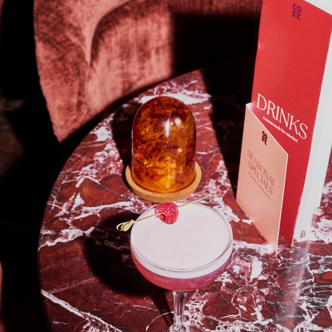 Artfully Crafted Cocktail Presentation A pink cocktail garnished with a raspberry sits on a marble table beside a glowing amber dome and a drinks menu.